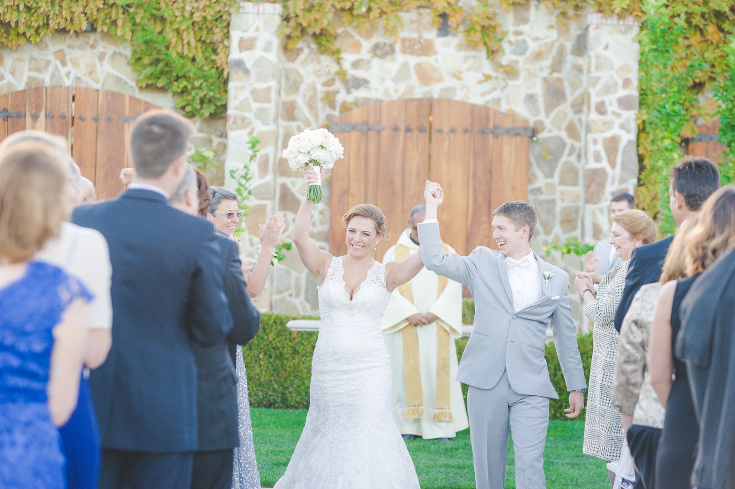 A couple celebrates joyfully during an outdoor wedding ceremony with guests gathered on a lawn by a stone wall.