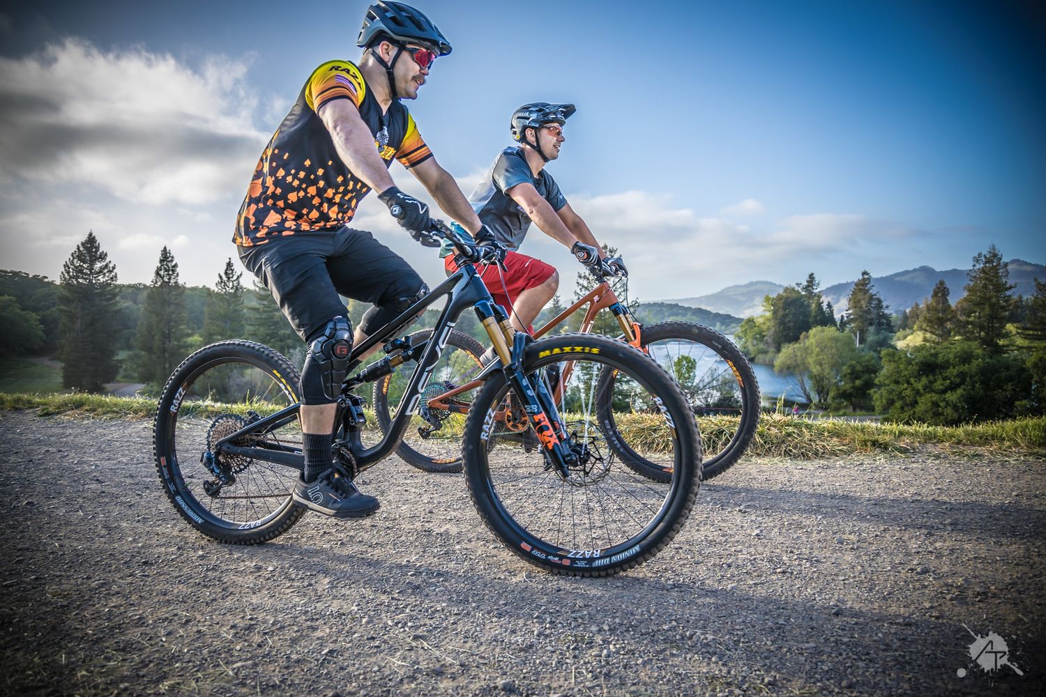 Two mountain bikers cycling together on a gravel path surrounded by mountain scenery and evergreen trees.
