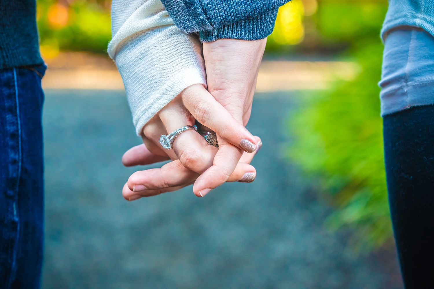 Close-up of two people holding hands with engagement rings visible against a blurred green natural background.