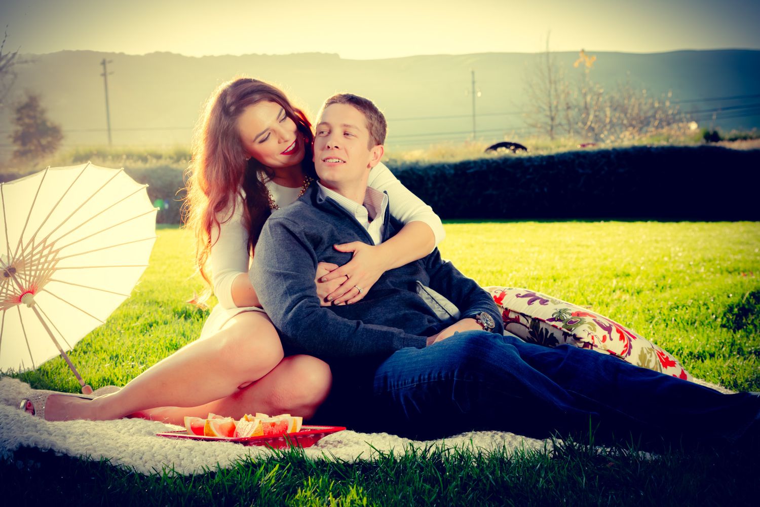 A romantic couple lounges on a grassy hillside with a white parasol on a sunny afternoon.