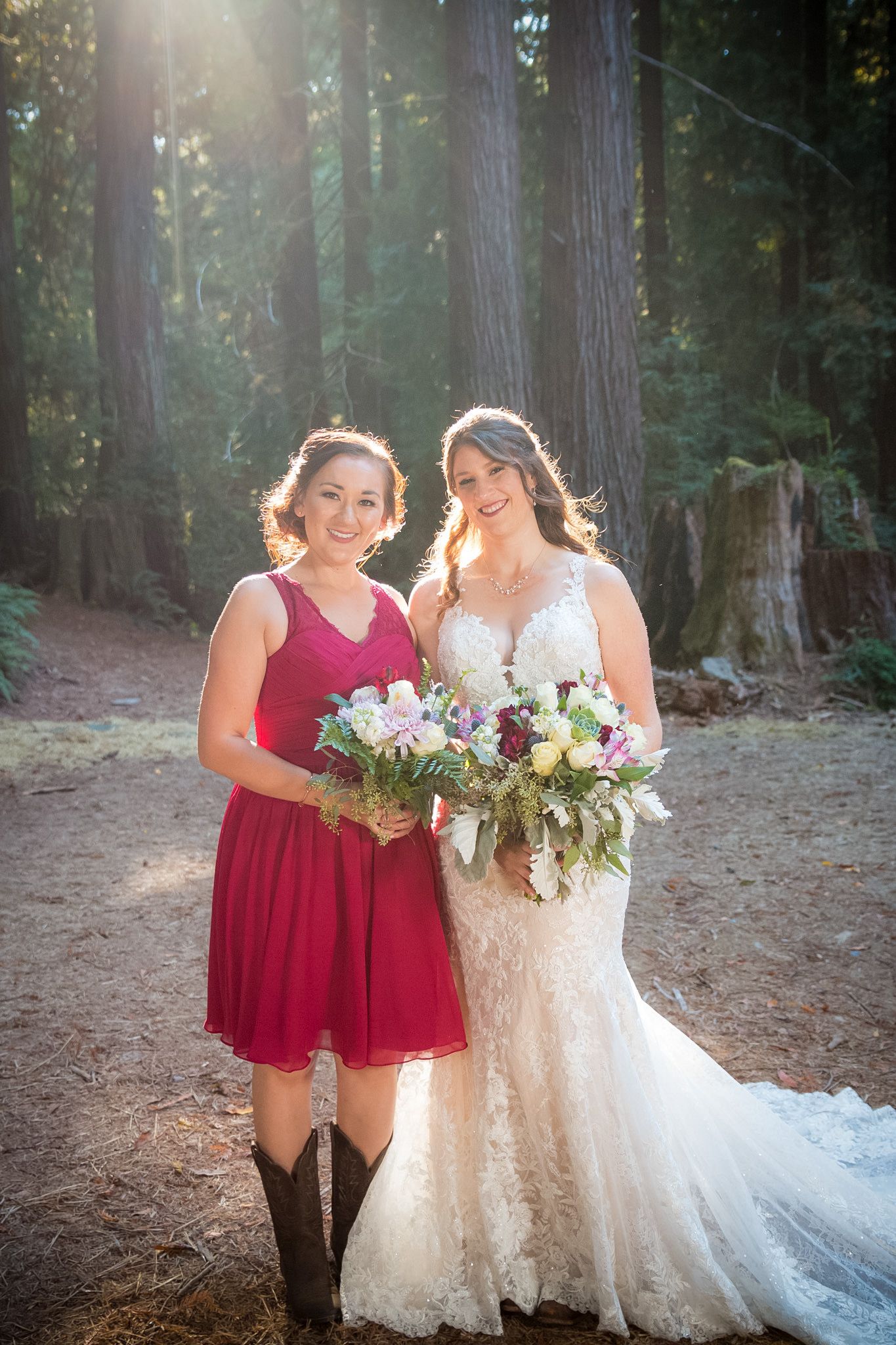 Bridesmaid in cowboy boots and bride pose with bouquets on forest trail.