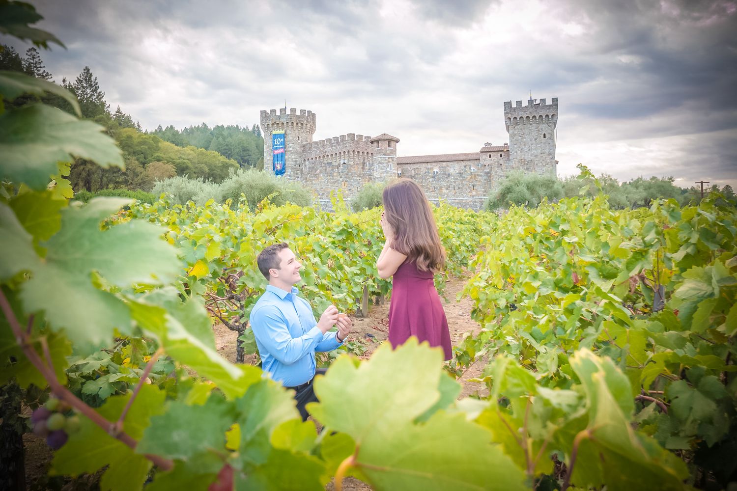 A romantic proposal moment in a vineyard with a castle in the background under cloudy skies.