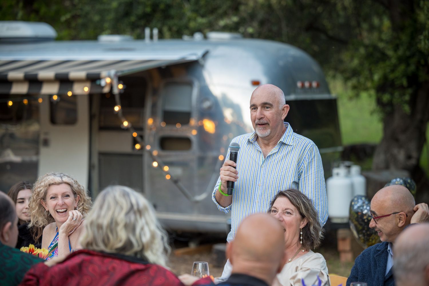 An outdoor dinner event with guests gathered around tables beneath string lights and an airstream trailer.