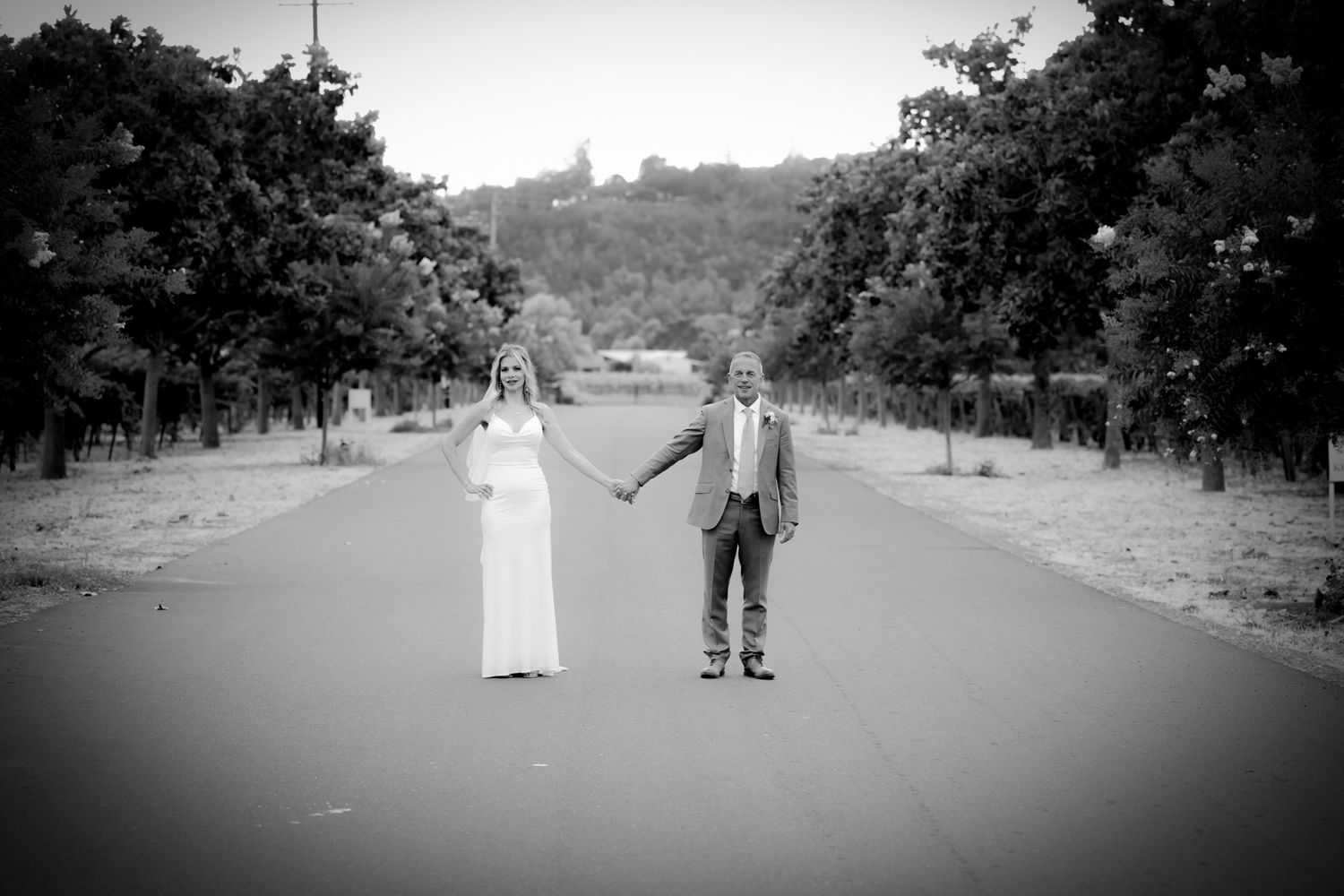 Black and white photo of couple holding hands on tree-lined road.