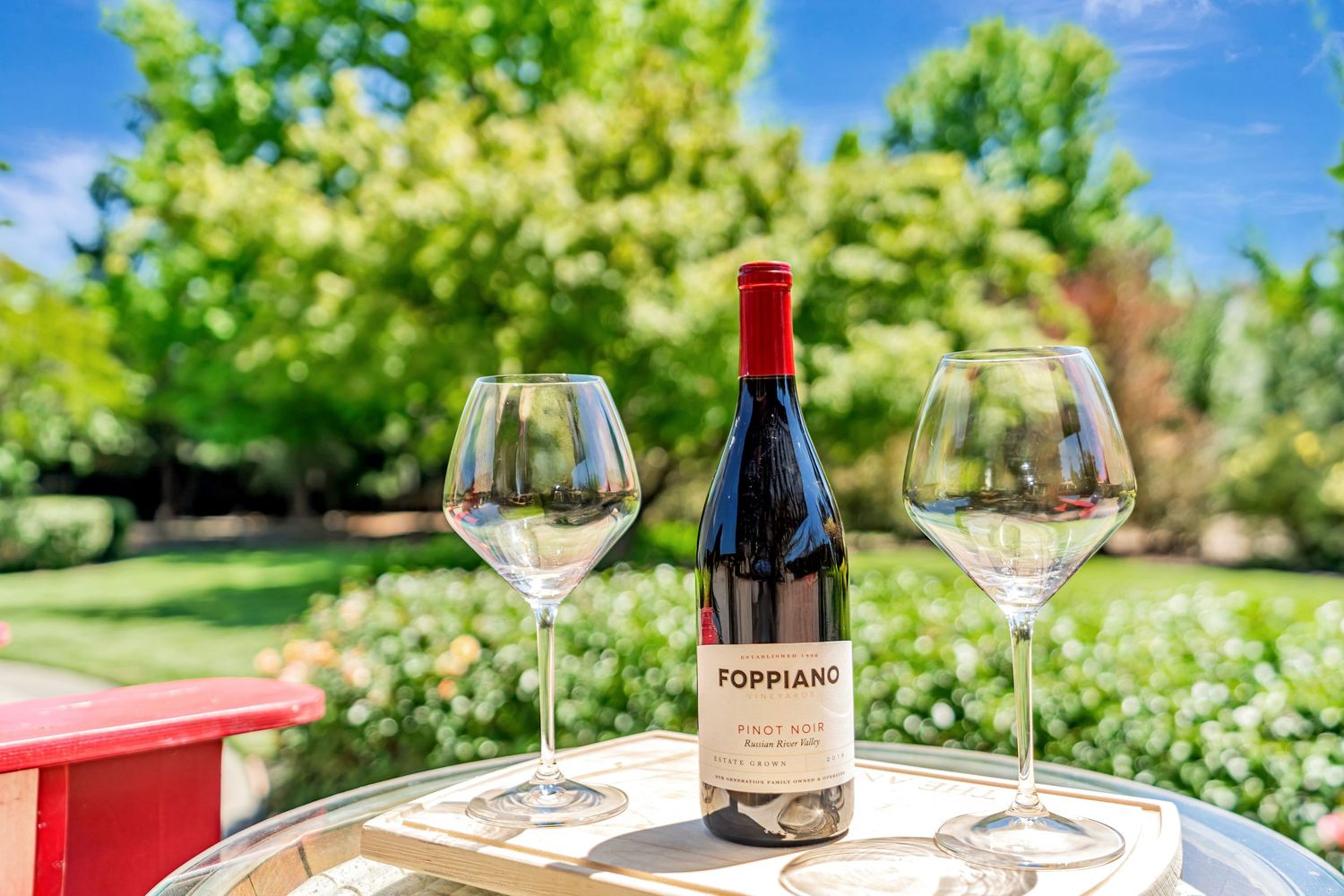 Wine bottle and glasses set on outdoor table with lush garden backdrop.