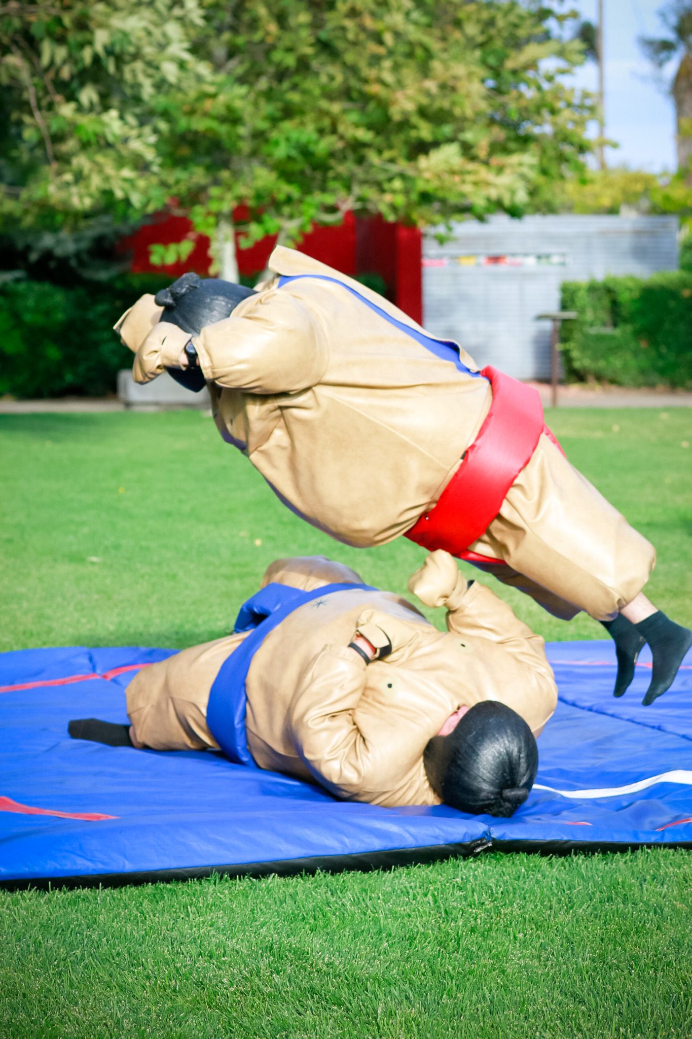 Two people in sumo wrestling suits playfully engaged in a match on a blue mat outdoors.
