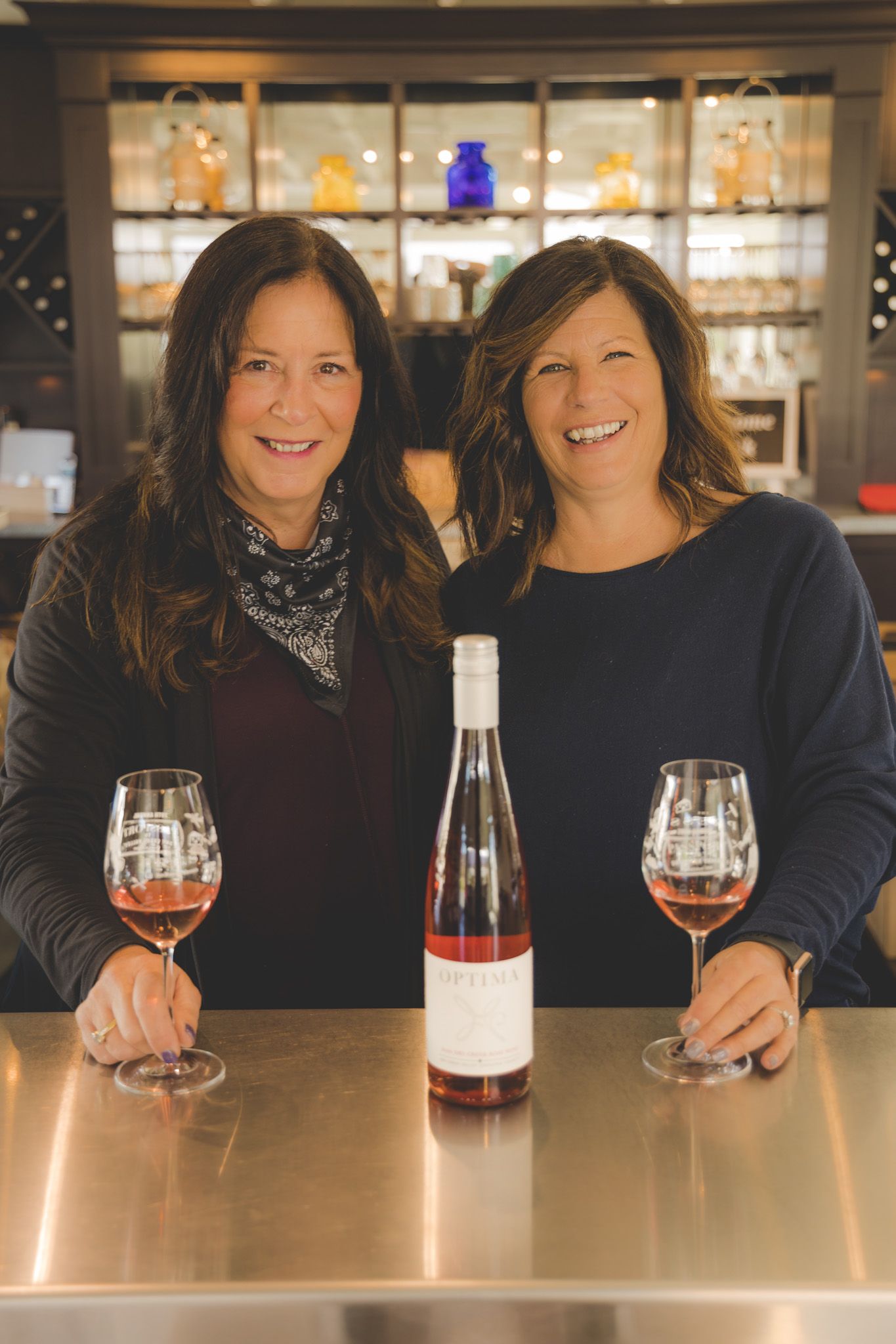 Two wine enthusiasts enjoying glasses of rosé at a tasting bar with bottles displayed behind them.