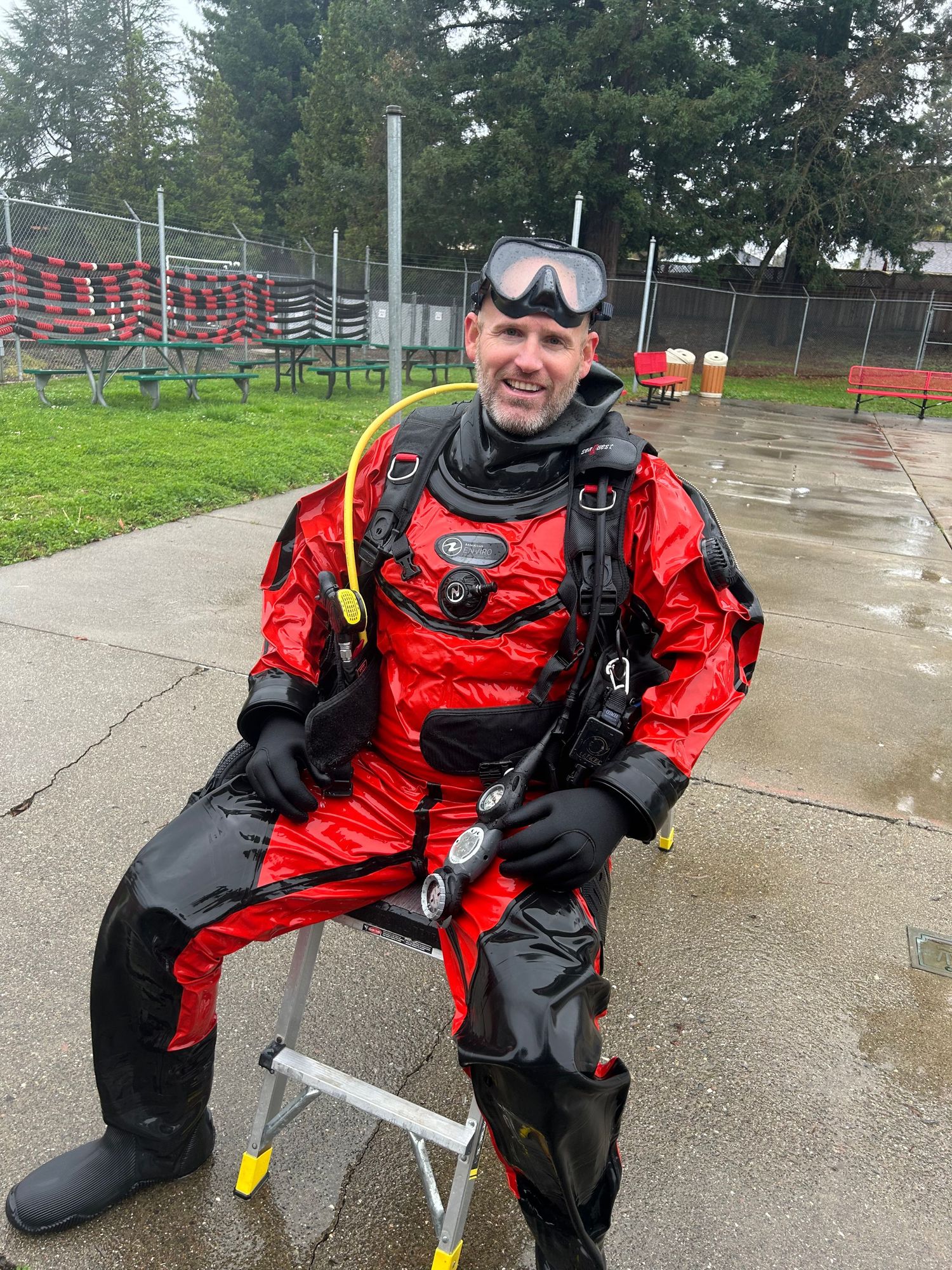 Diver in bright red drysuit sitting on metal stool with diving gear.