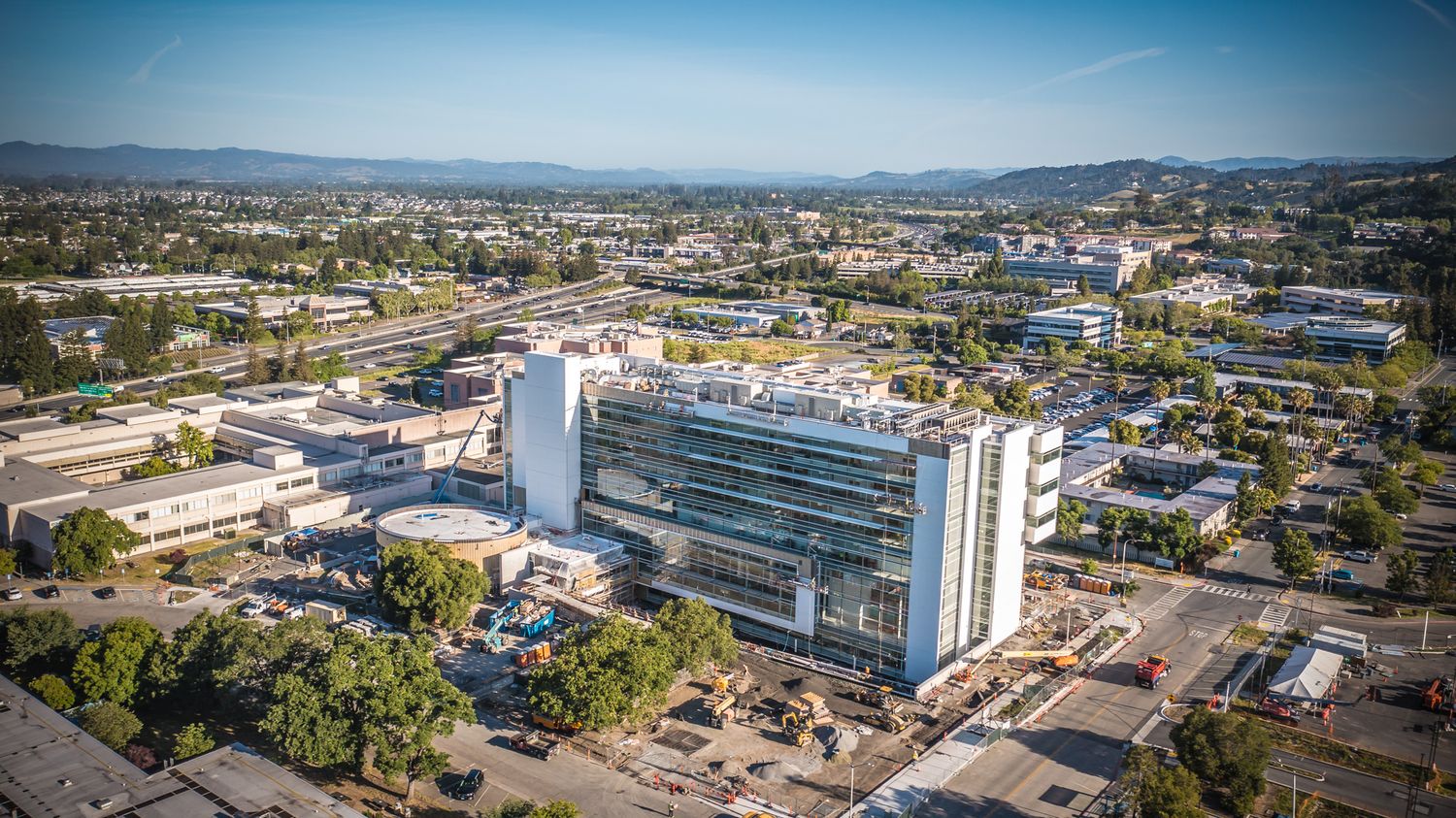 Modern glass office building with multiple stories surrounded by parking lots and mountains in the background.