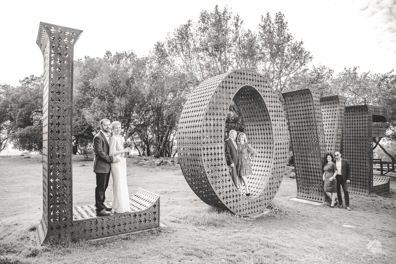 Black and white photo of couples posing with giant decorative LOVE letters in an outdoor garden setting.