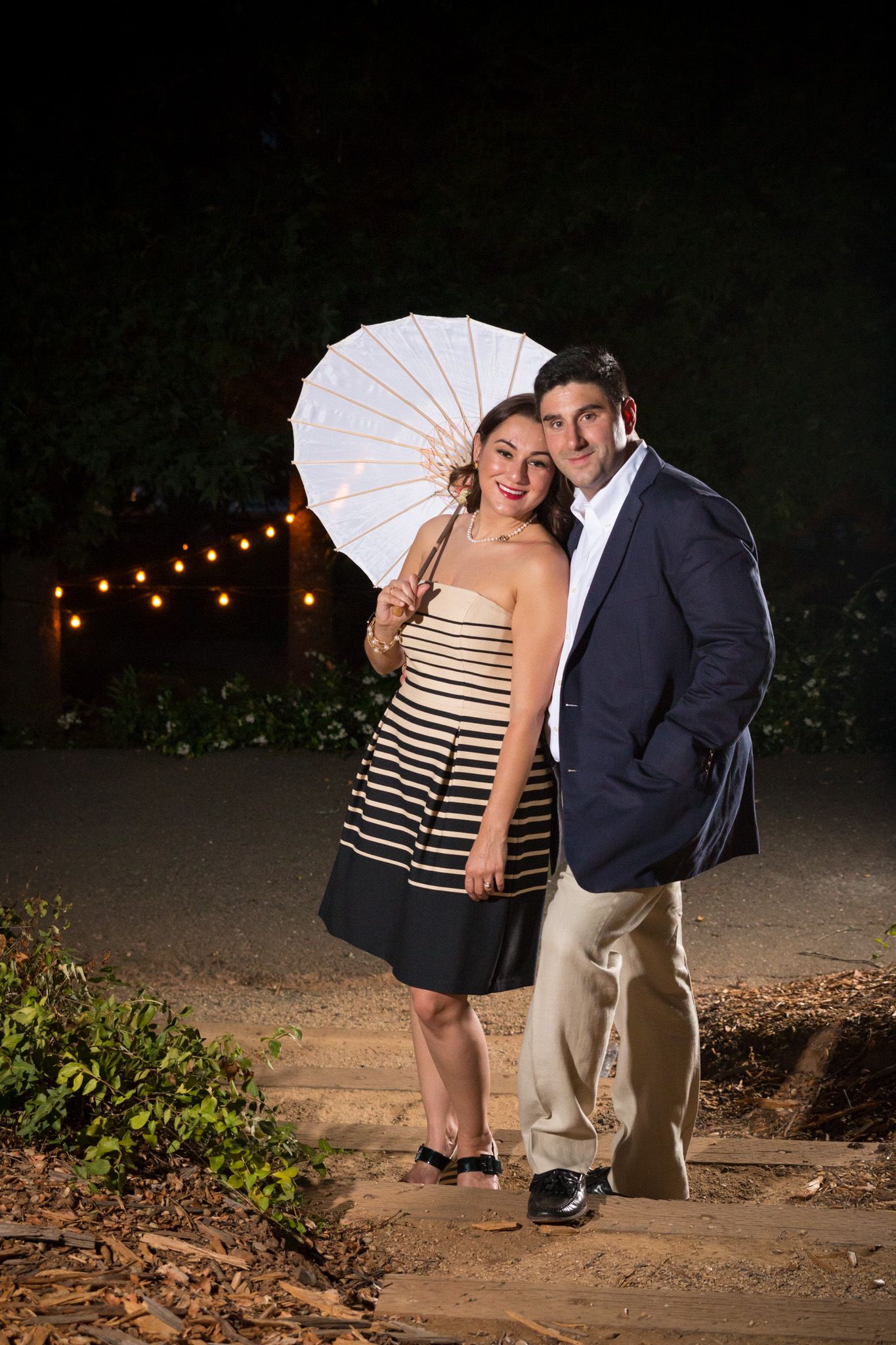 Romantic moment under string lights as couple poses with white parasol at nighttime.