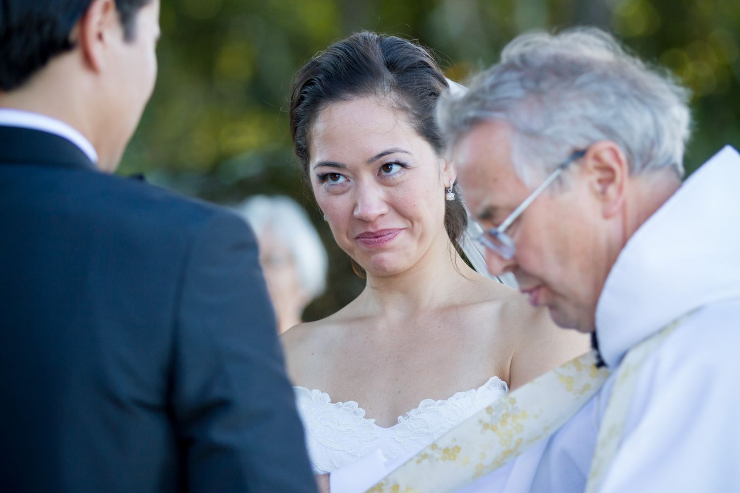 A wedding ceremony with a bride in white dress listening to the officiant outdoors.