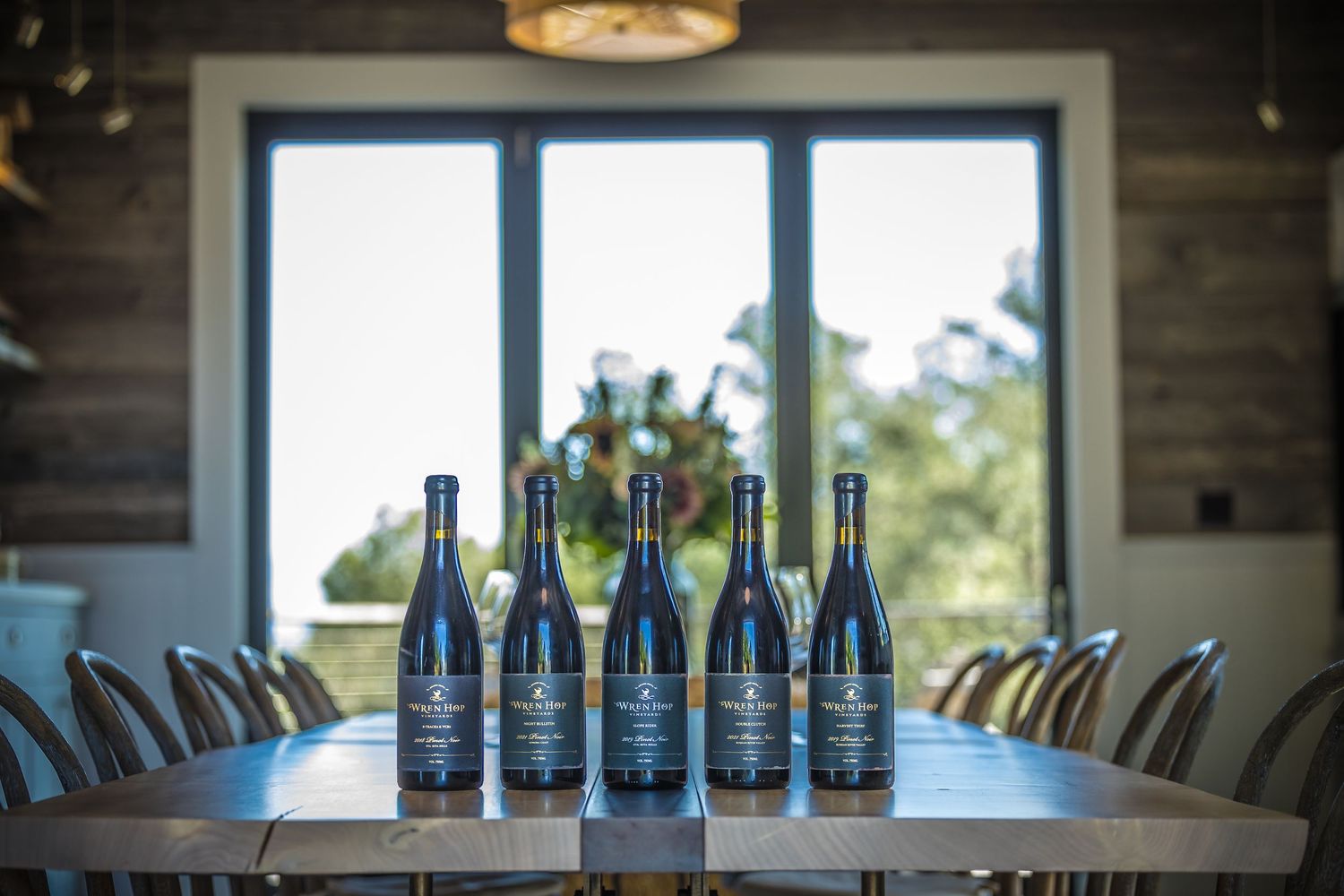 Row of wine bottles displayed on long dining table with chairs and window view.
