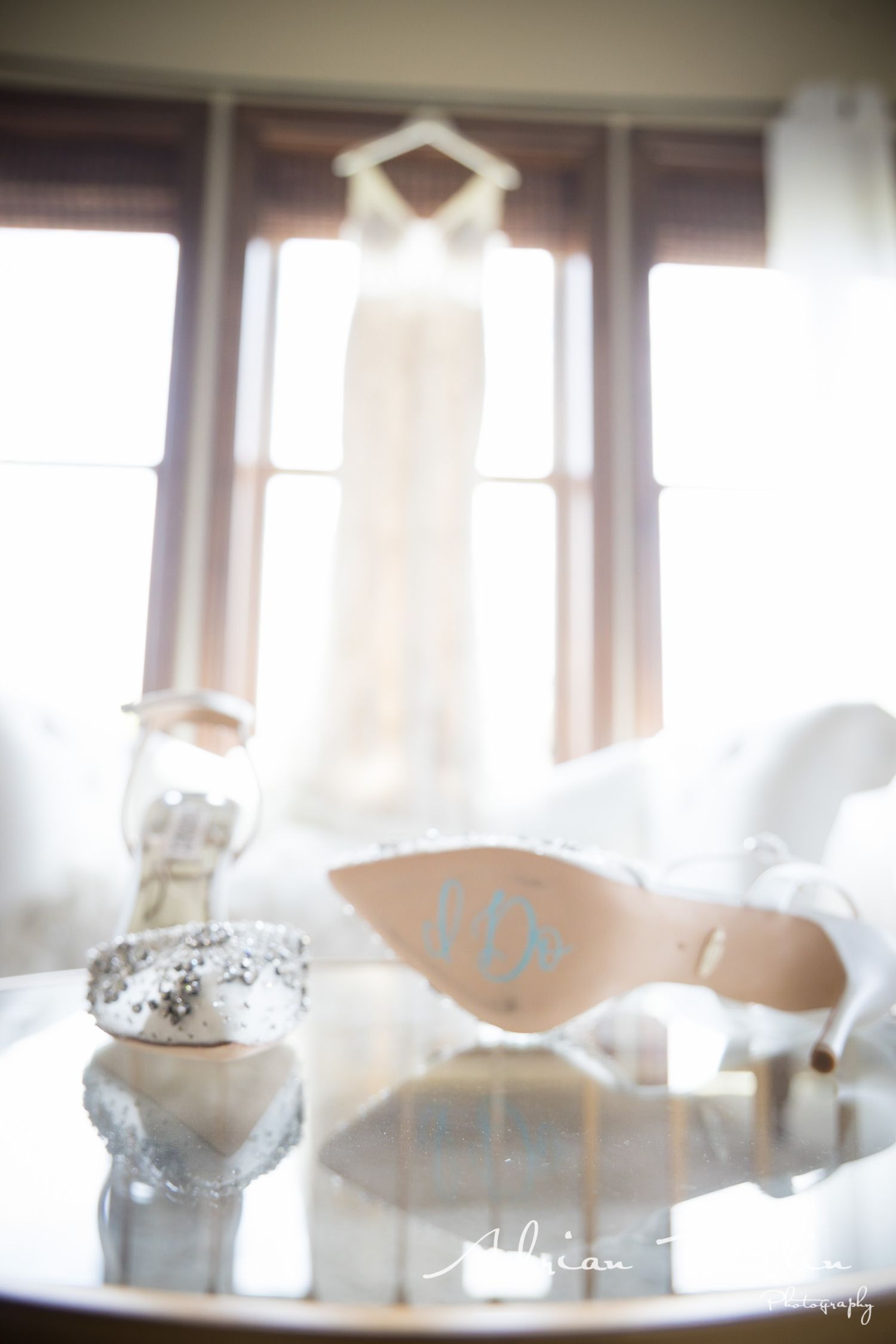 A white wedding dress hangs in silhouette against a bright window with bridal shoes below.
