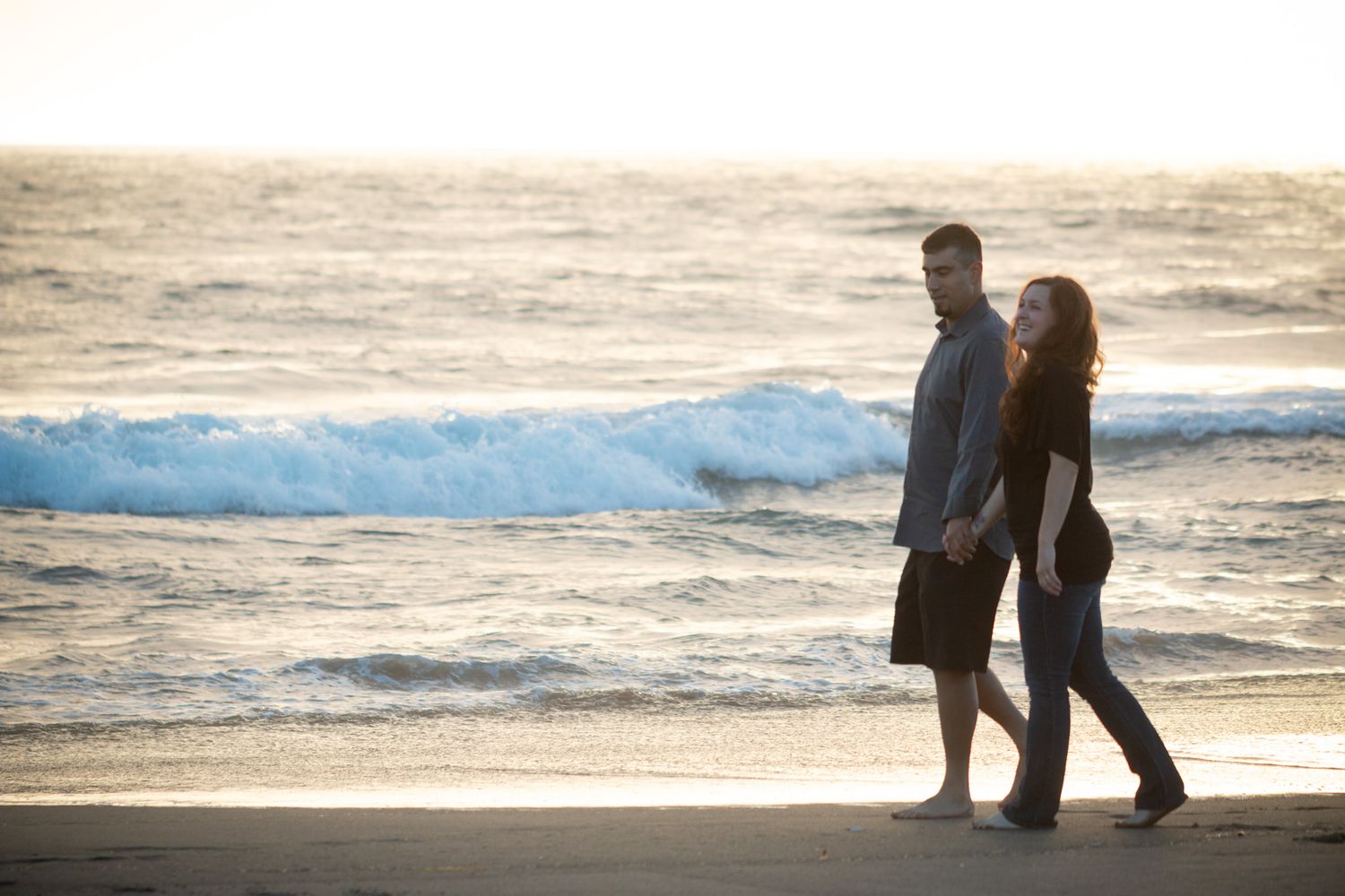 Couple walking together along beach with waves and sunset in background.