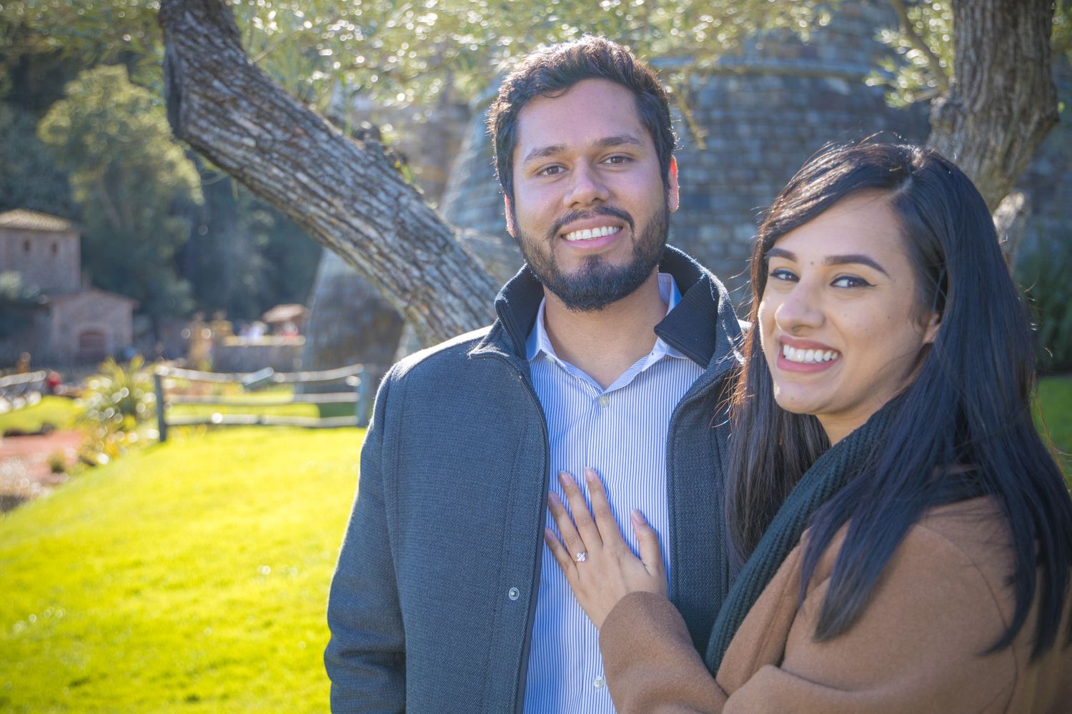 A couple stands together on a sunny lawn with trees in the background.