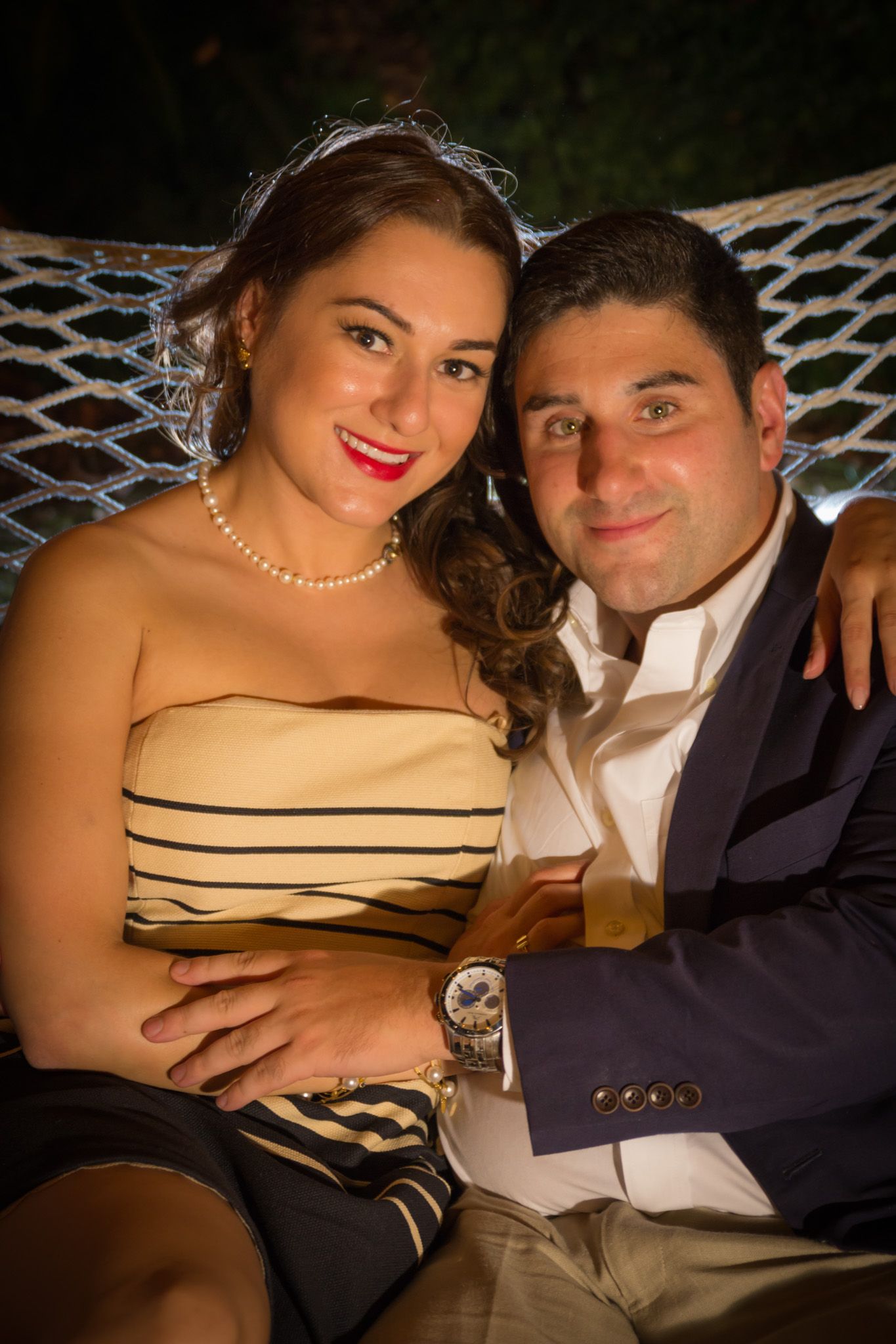 Couple sits together on wicker furniture at formal event, wearing elegant evening attire.