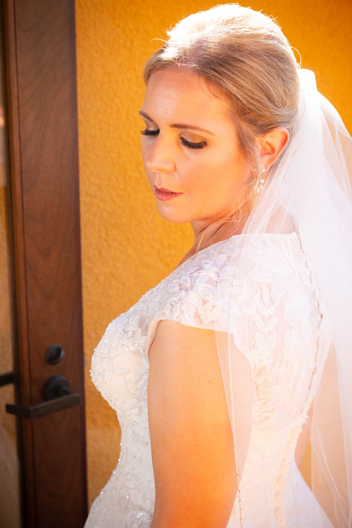 Side profile of bride in lace gown with flowing veil against orange wall.