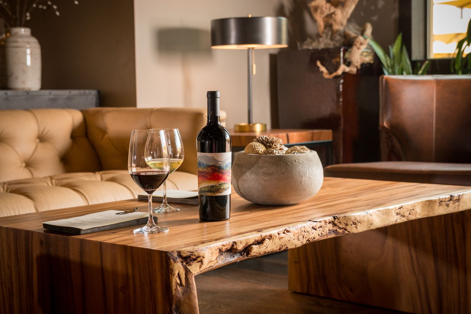 Wine bottle and glass on rustic live edge wooden table with modern table lamp and leather seating.