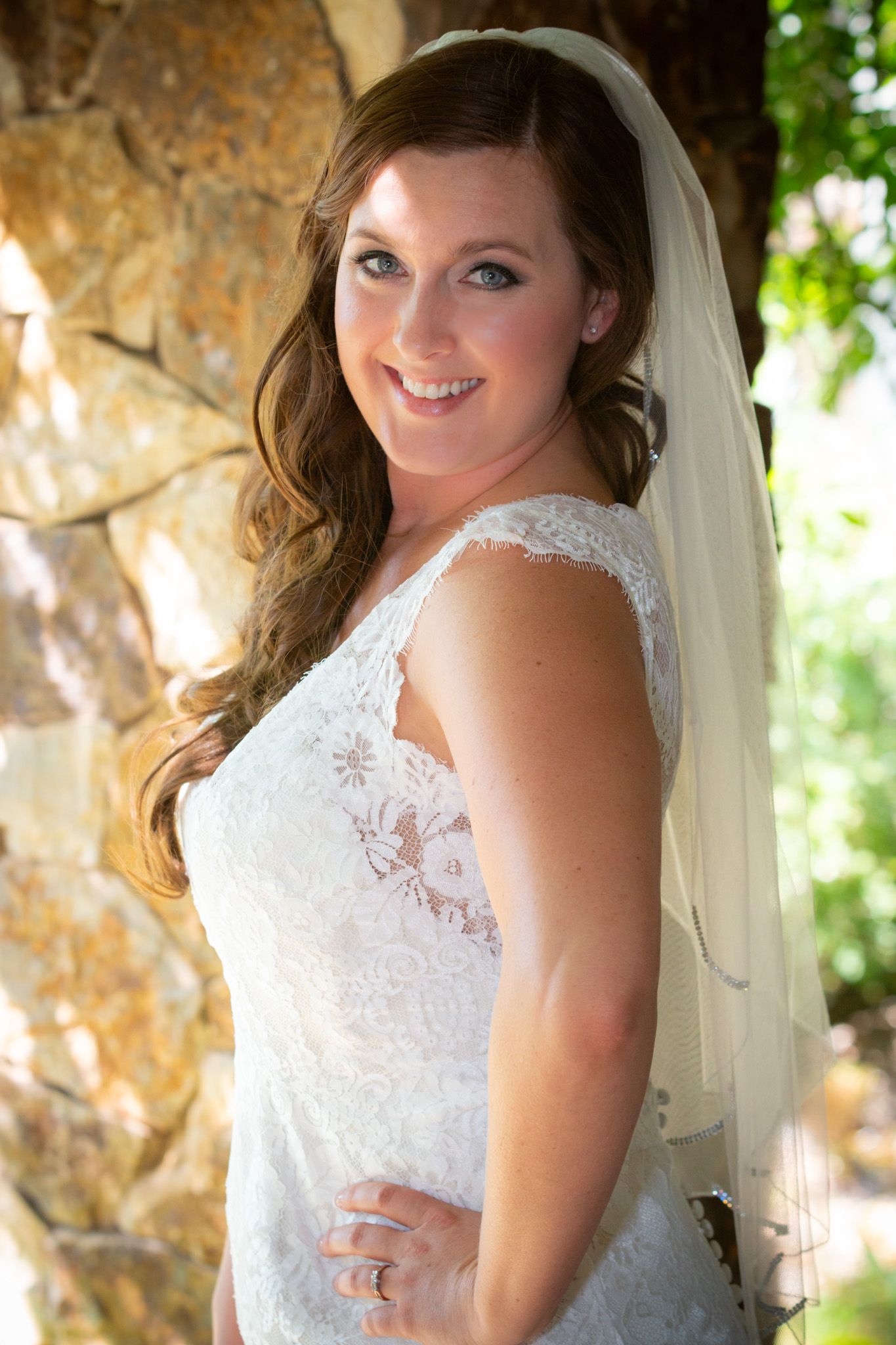 A beautiful bride in a lace wedding dress poses against a stone wall.