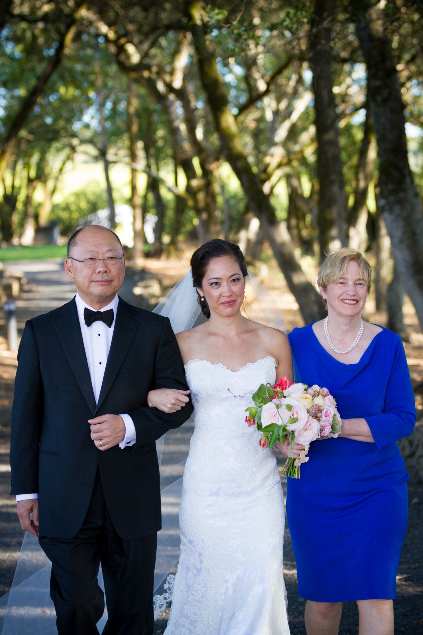 Wedding procession walking down tree-lined path with couple and attendant in elegant formal attire.
