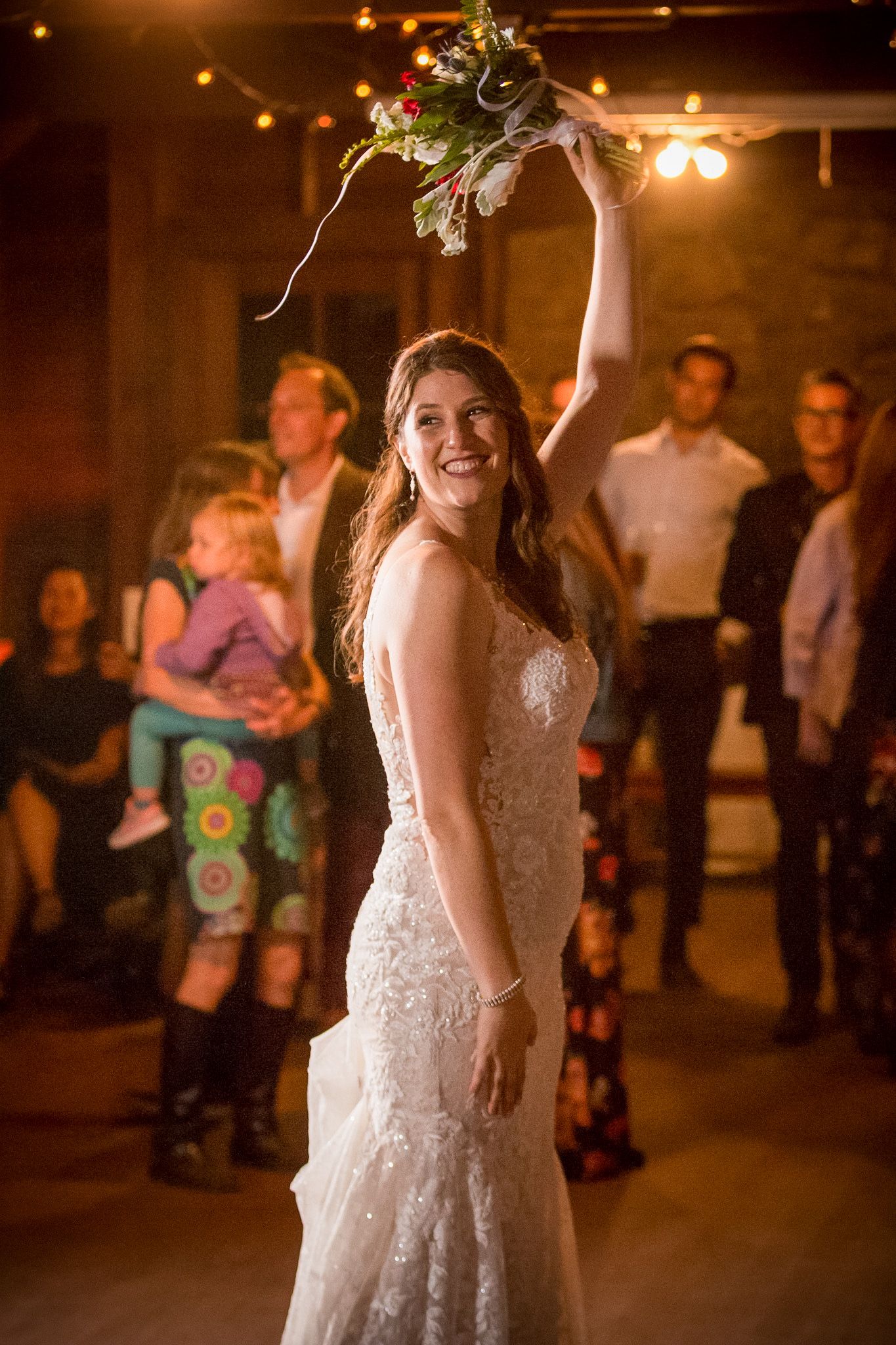 A bride in a lace wedding dress holds up her bouquet during a reception celebration.