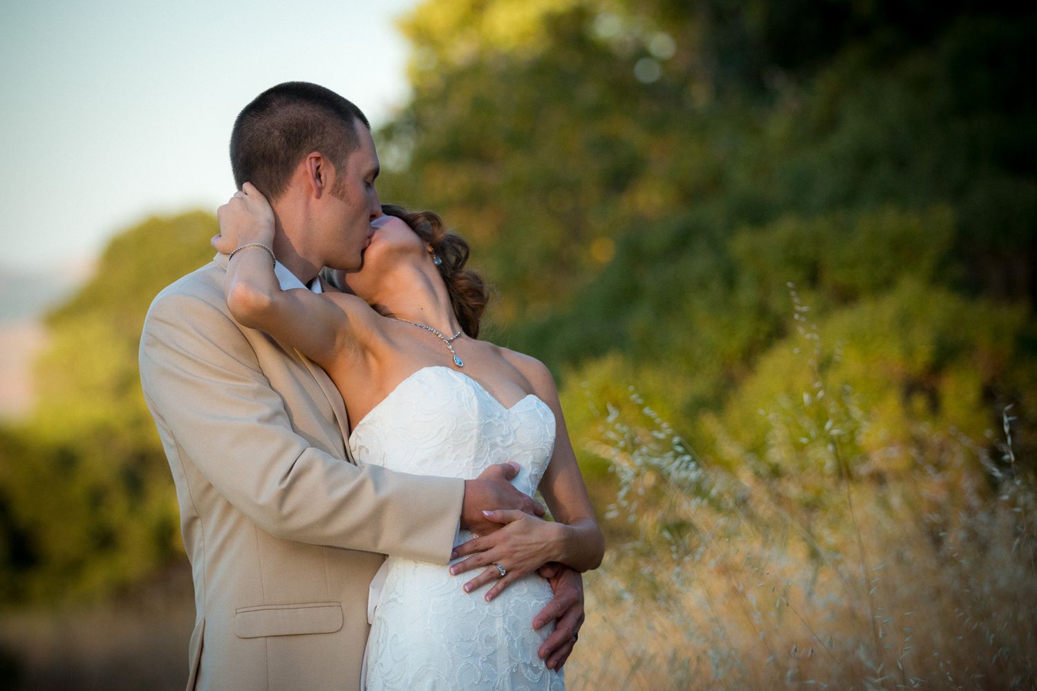 Romantic wedding couple embraces outdoors in golden sunset light among tall dried grasses in garden setting.