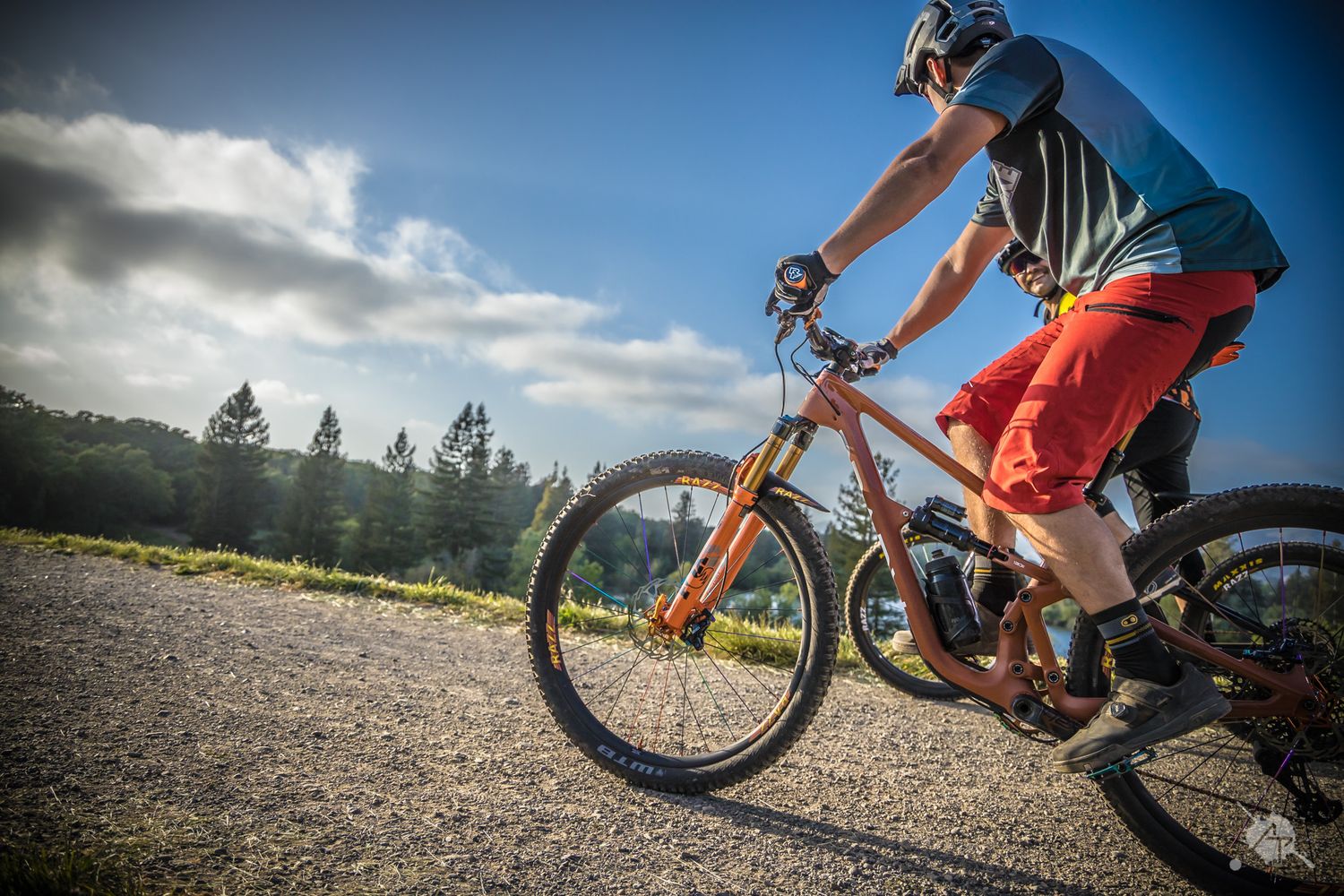 Mountain biker rides on a scenic trail with orange shorts and blue shirt against cloudy sky and mountain backdrop.