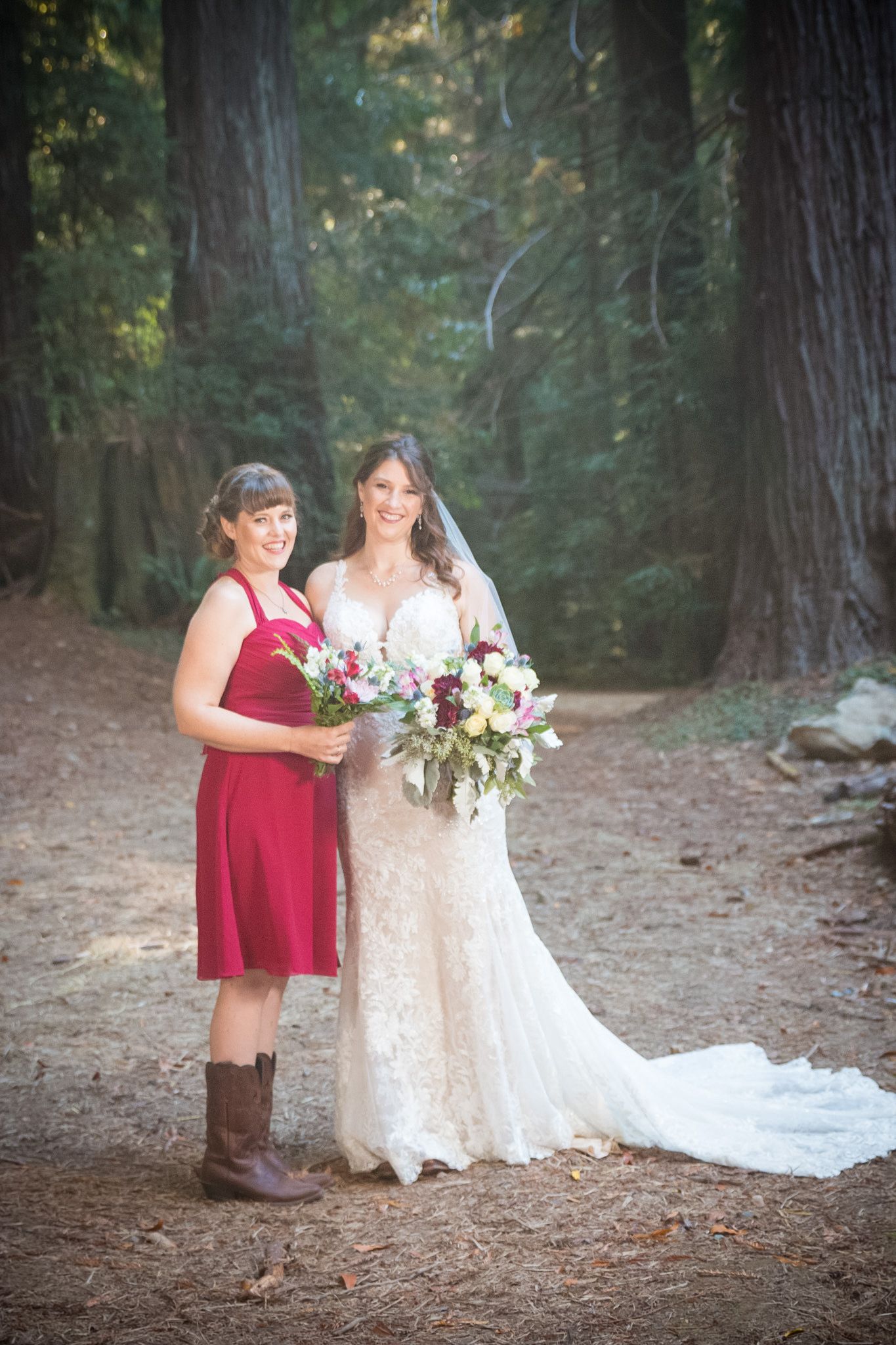 Bride and bridesmaid holding floral bouquets pose together on forest trail among redwood trees.