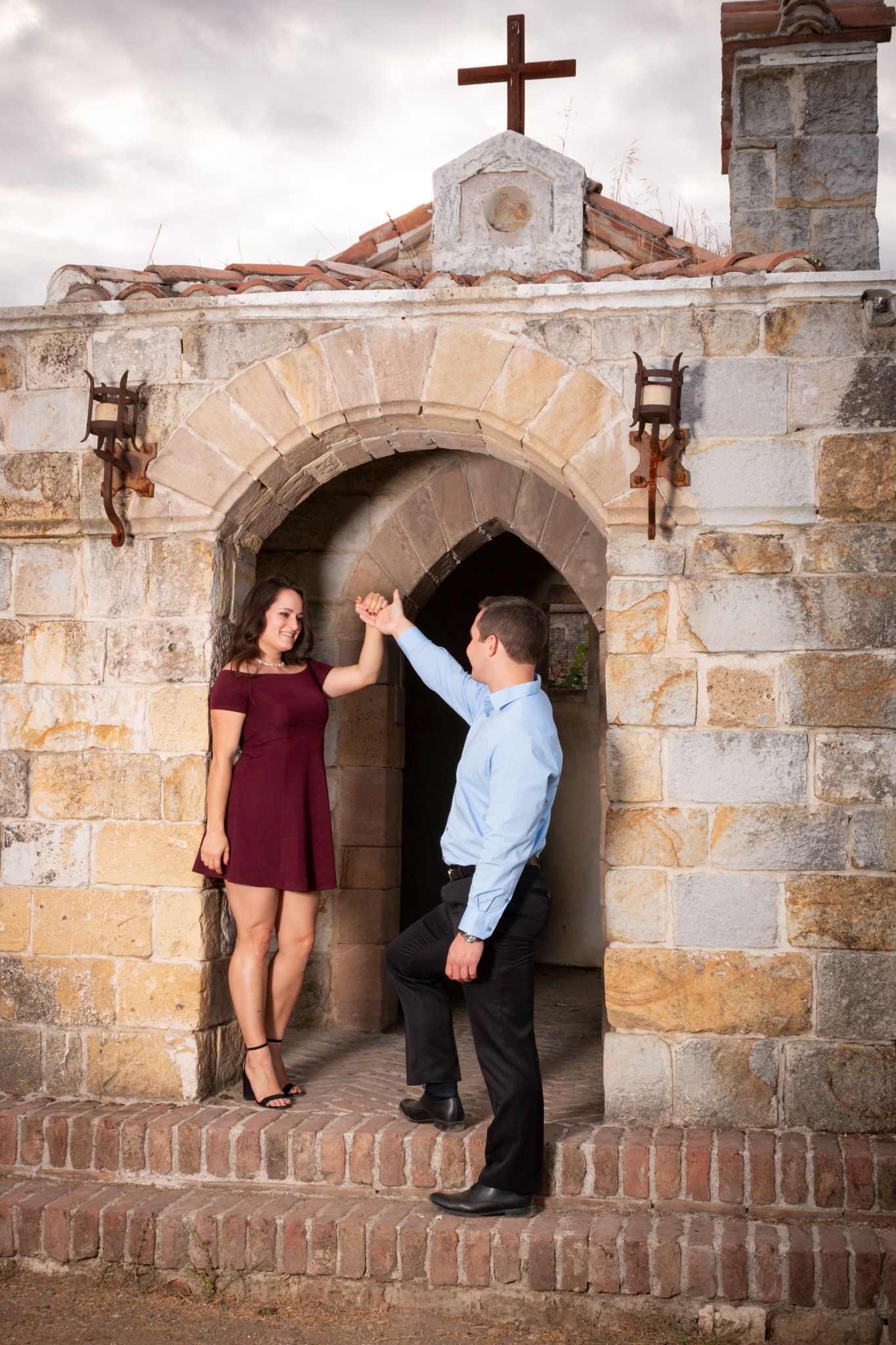 Couple dancing under stone archway with cross at mission-style building.