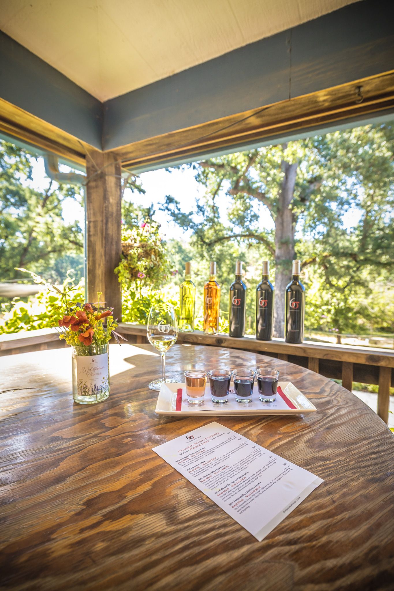 Wine tasting setup with bottles, glasses and menu on rustic table overlooking scenic outdoor view.
