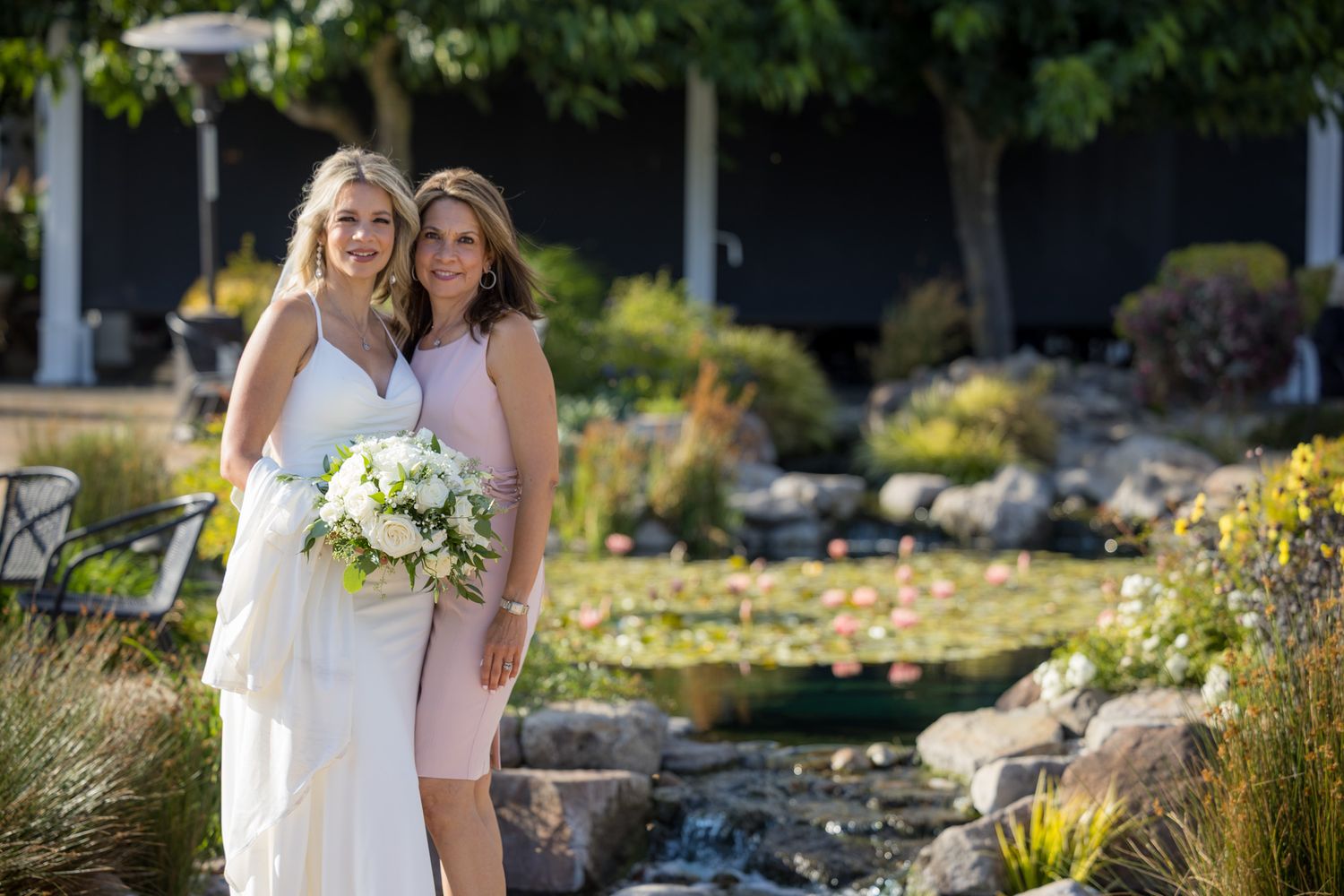 Two people in white dresses standing by water feature in garden setting.