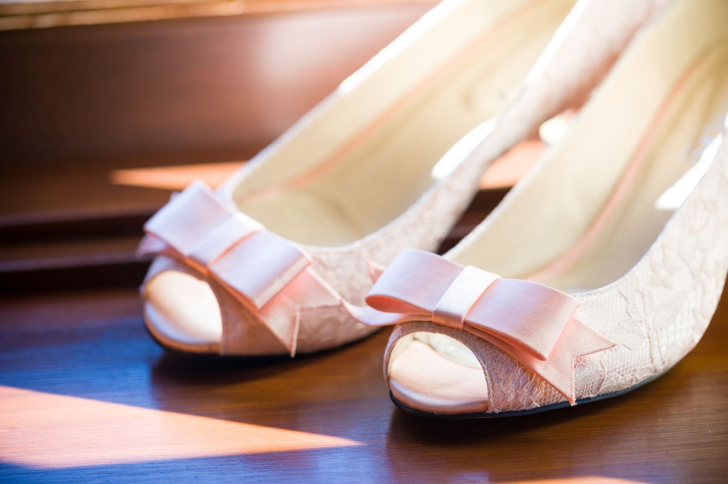 White lace bridal peep-toe shoes with pink bows sit elegantly on a wooden surface in soft lighting.