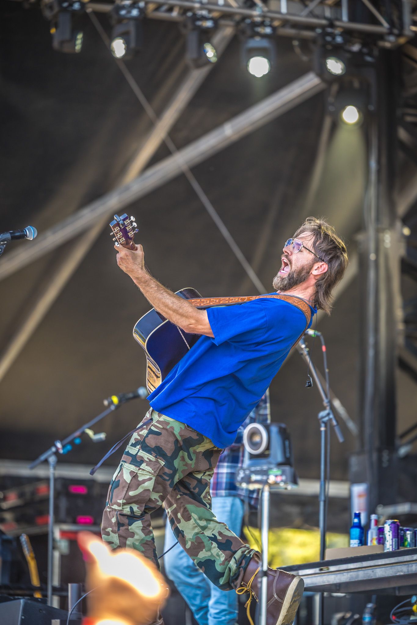 Musician performing energetically with acoustic guitar on stage with dramatic lighting and metal rigging overhead.