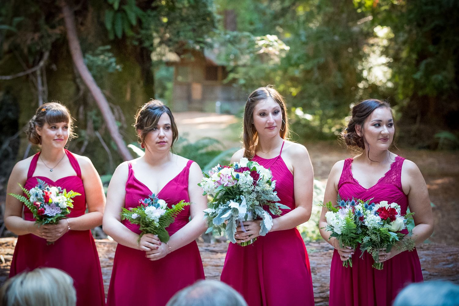 Four bridesmaids in matching magenta dresses holding floral bouquets at outdoor ceremony.