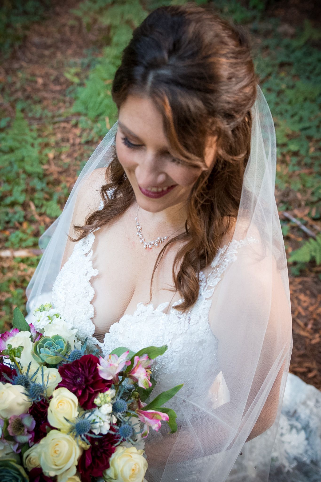 A bride in a lace wedding dress holds a colorful bouquet with red and cream roses in a forest setting.