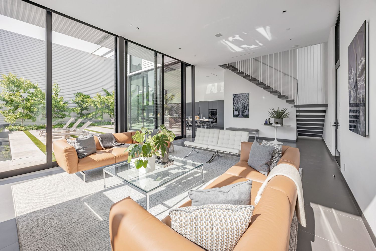 Bright modern living room with tan leather chairs, floating staircase, and floor-to-ceiling windows.