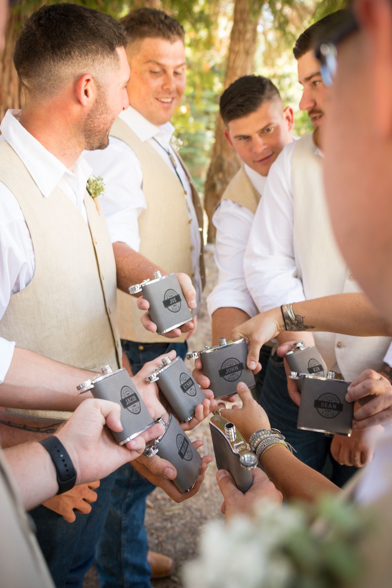Groomsmen in light colored vests gather to look at matching koozies outdoors.
