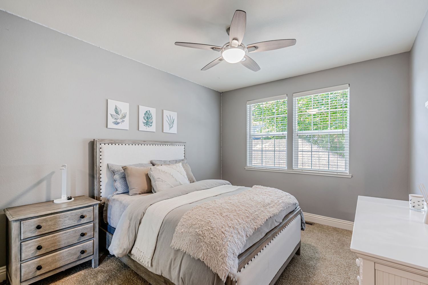 Peaceful bedroom with gray walls featuring white artwork, ceiling fan, and light beige upholstered headboard.
