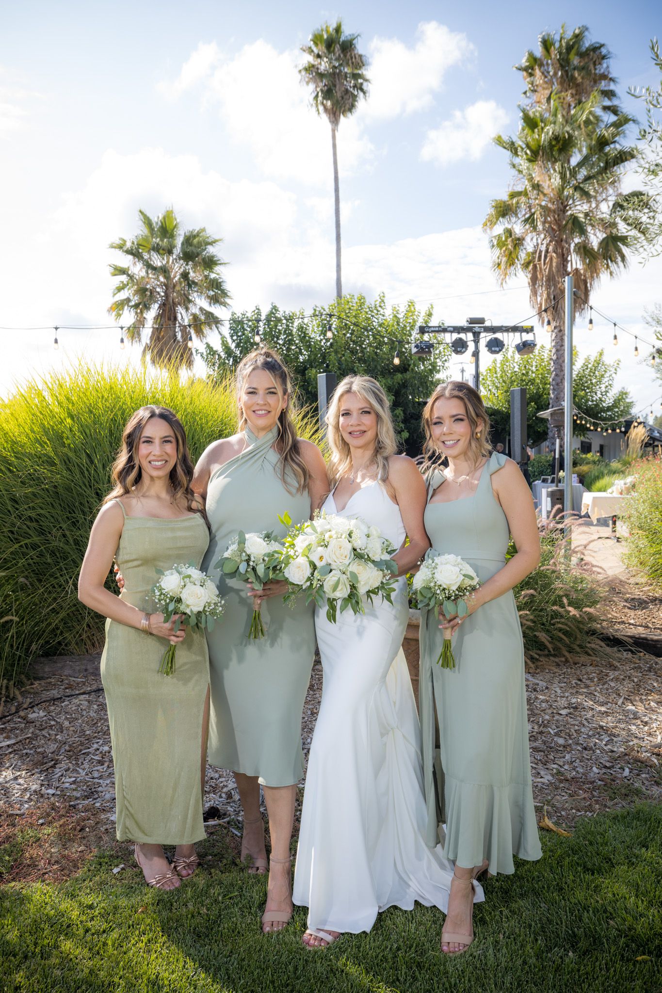 Bridal party in sage green dresses poses with white bouquets against palm trees and garden.