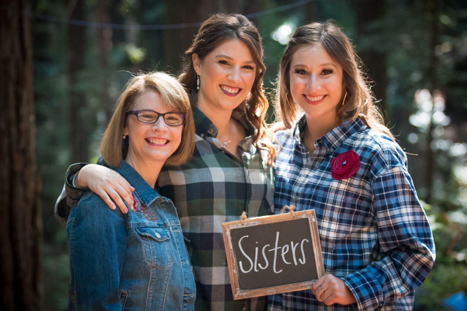 Three women in plaid shirts and denim holding a chalkboard sign reading 'Sisters' in an outdoor forest setting.