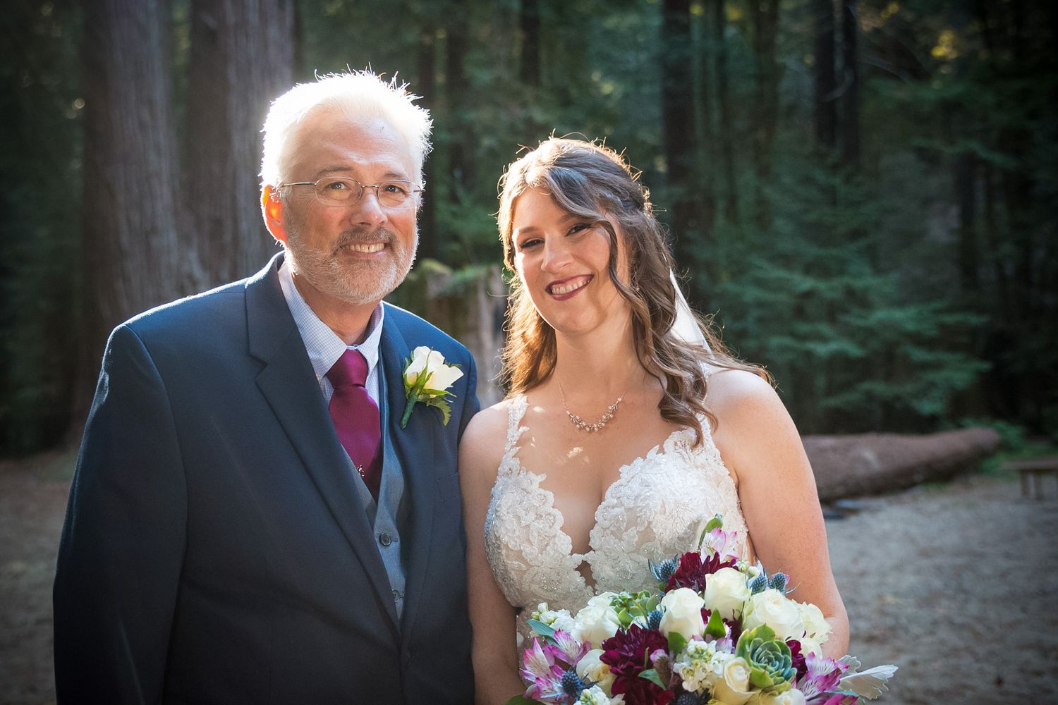 Wedding portrait of a bride and older gentleman in formal attire posing in a forest setting.