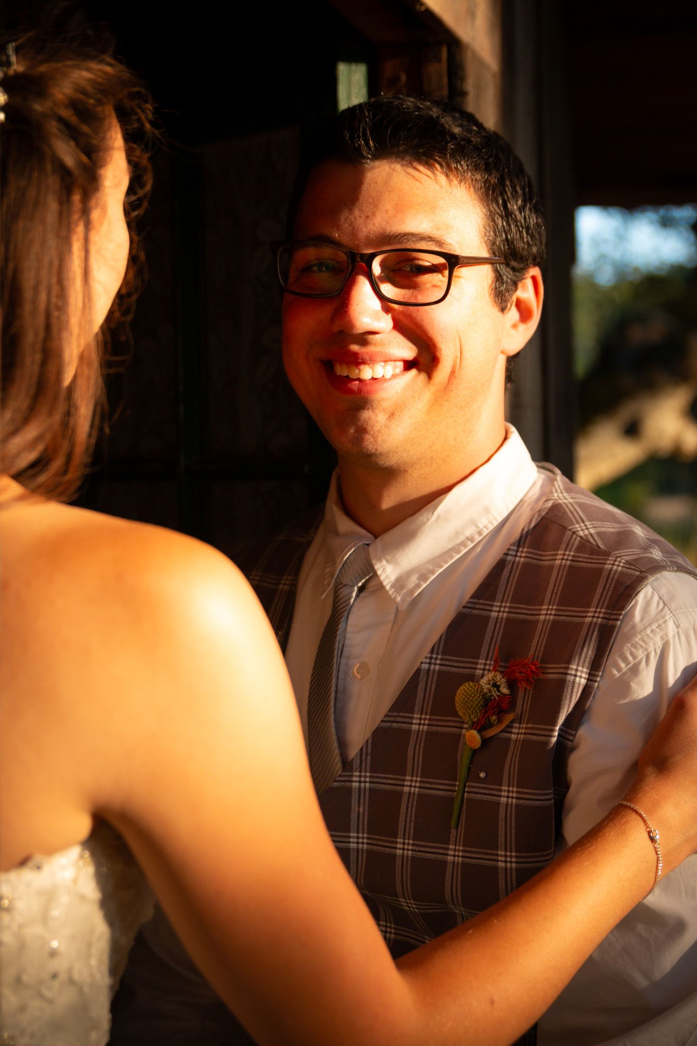 A person wearing glasses and a plaid vest smiles while wearing a boutonniere at an evening wedding celebration.