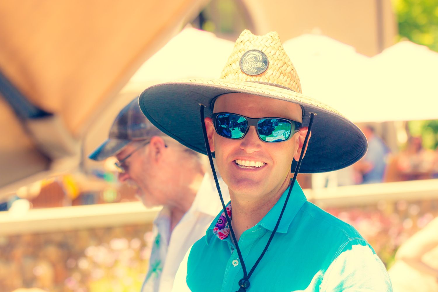 Person wearing a wide-brimmed hat, sunglasses and teal shirt smiling outdoors on a sunny day.