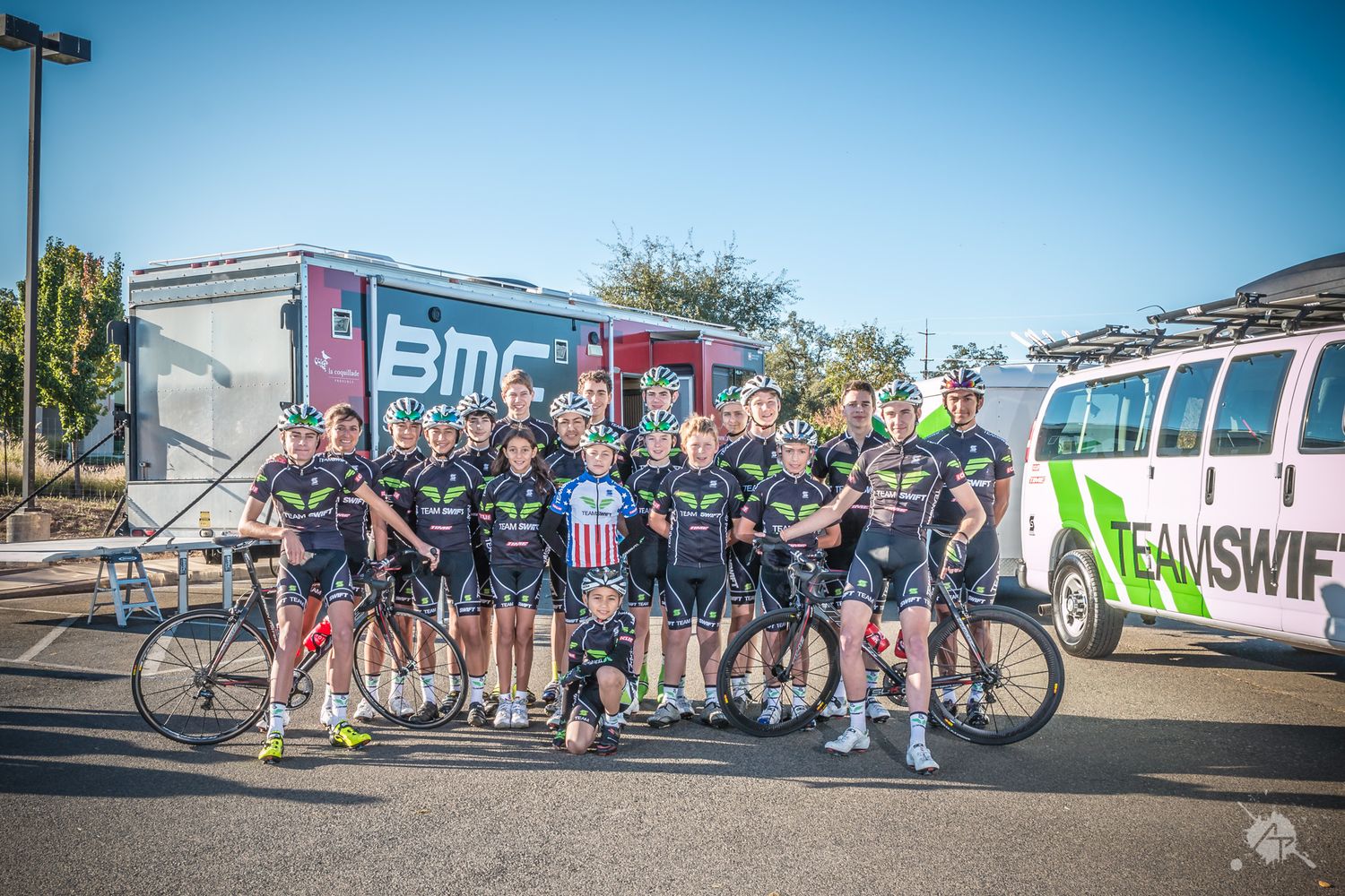 BMC cycling team poses together with their bikes and support vehicles in a parking lot.
