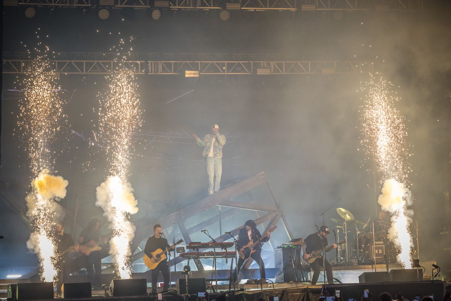 Concert stage performance with dramatic pyrotechnic effects and spotlights illuminating musicians through atmospheric haze.