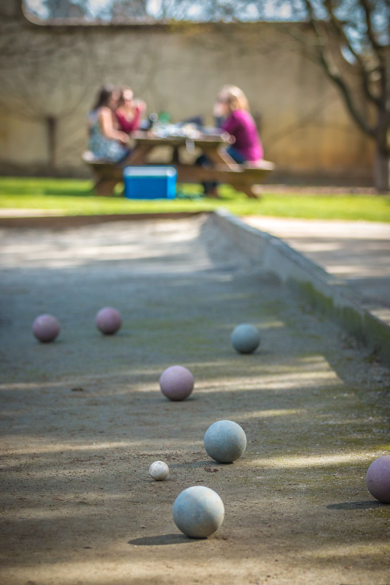 Bocce balls scattered on a sandy court with players sitting at a picnic table in the background.