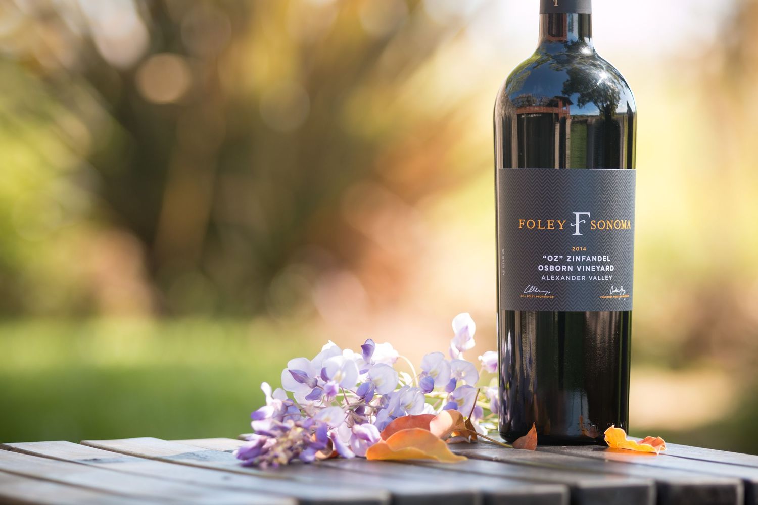 Wine bottle with purple flowers on wooden table in outdoor setting.