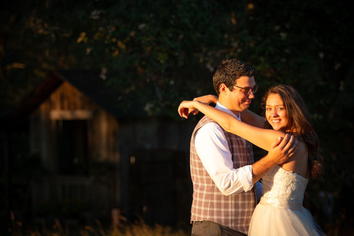 A couple embraces in romantic golden sunlight with a rustic barn in the background.