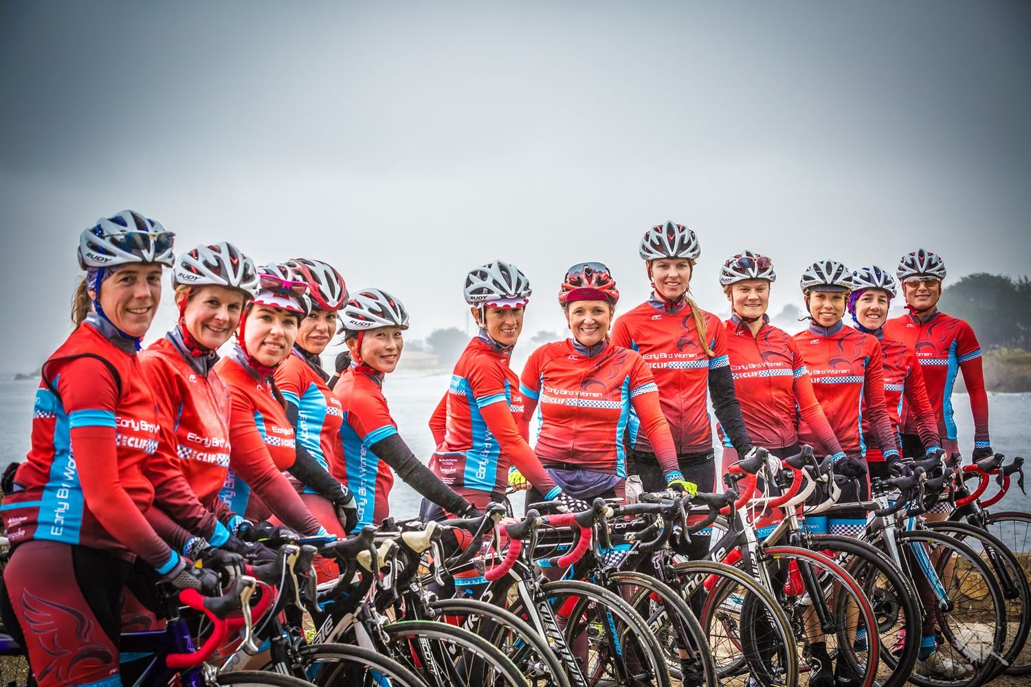 A group of cyclists in matching red and blue uniforms pose with their road bikes against a grey sky background.